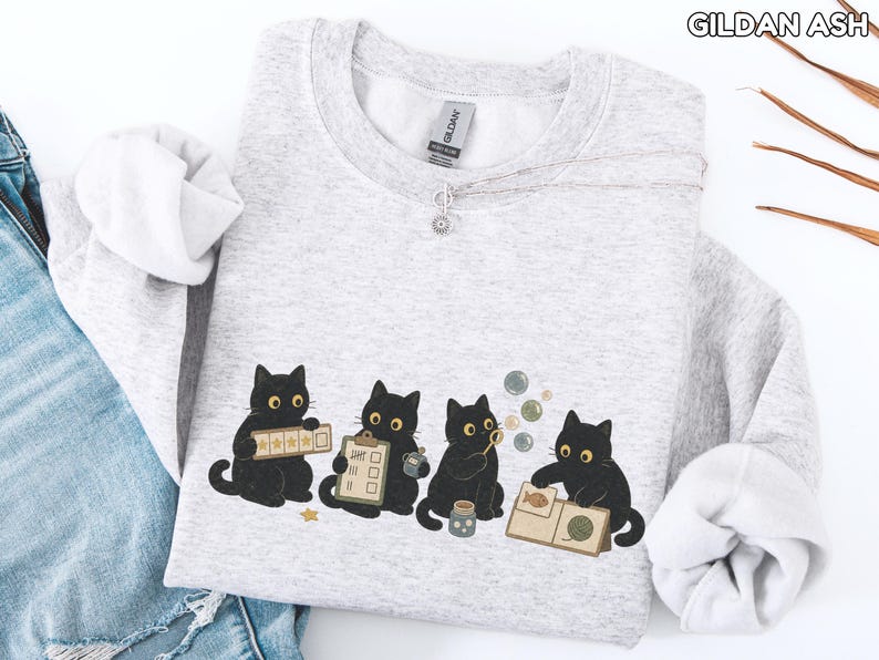 May include: A light gray Gildan Ash crewneck sweatshirt featuring a graphic of four black cats. One cat holds a five-star rating card, another a clipboard, the third blows bubbles, and the fourth examines a book with a fish and yarn illustration.