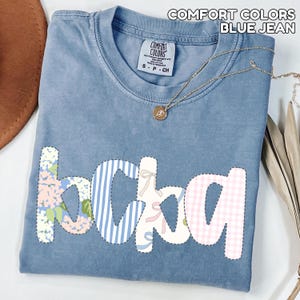 May include: Blue jean Comfort Colors t-shirt with the word "bckag" in a patchwork design. The letters feature floral, striped, and gingham patterns. A gold necklace with a pendant is draped across the shirt.