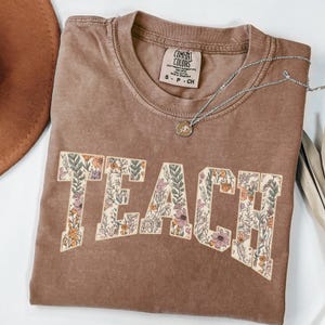 May include: A brown t-shirt with the word "TEACH" printed in white letters. The letters are filled with a floral pattern in shades of pink, orange, and green. The shirt is made of a soft, comfortable material.