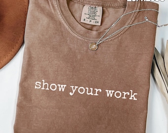 May include: Espresso-colored t-shirt with the text "show your work" in white. The shirt has a crew neck and a small tag near the collar. A silver necklace with a pendant is draped across the shirt. The words "COMFORT COLORS" and "ESPRESSO" are printed at the top.