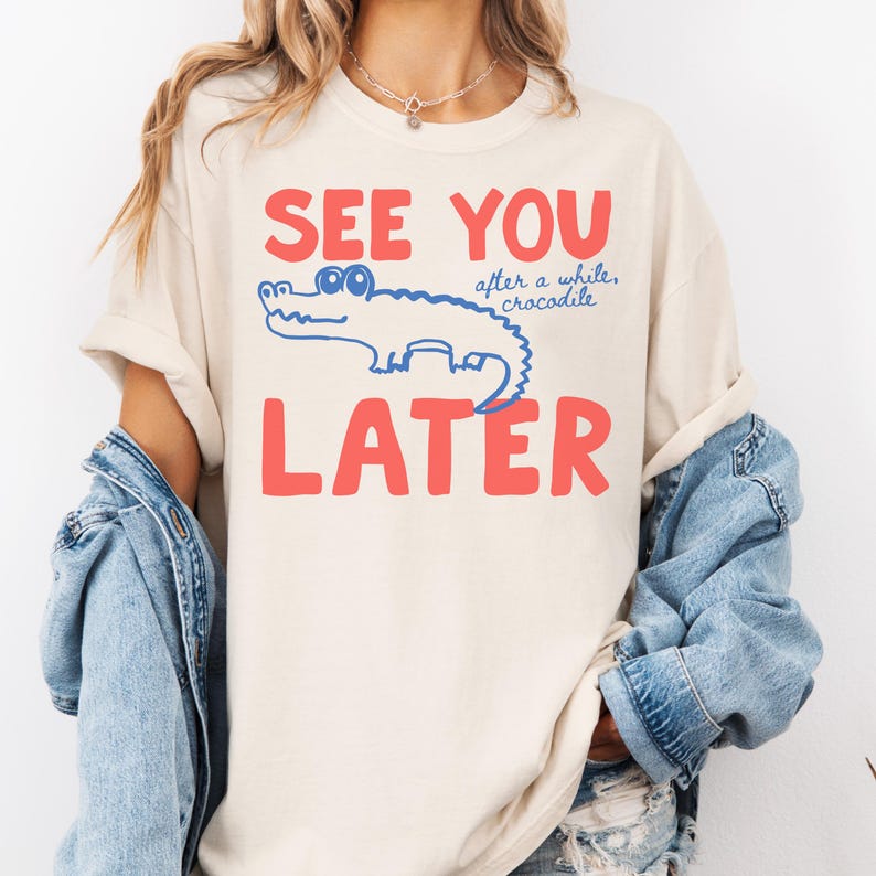 May include: A cream-coloured t-shirt featuring the words "SEE YOU LATER" in red block letters. A blue crocodile illustration is between the words. The phrase "after a while, crocodile" is above the crocodile.