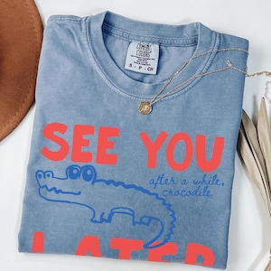 May include: A blue Comfort Colors t-shirt with the words "SEE YOU LATER" in red block letters. A blue cartoon crocodile is under the words. The phrase "after a while, crocodile" is written in blue script. A gold necklace is on the shirt.