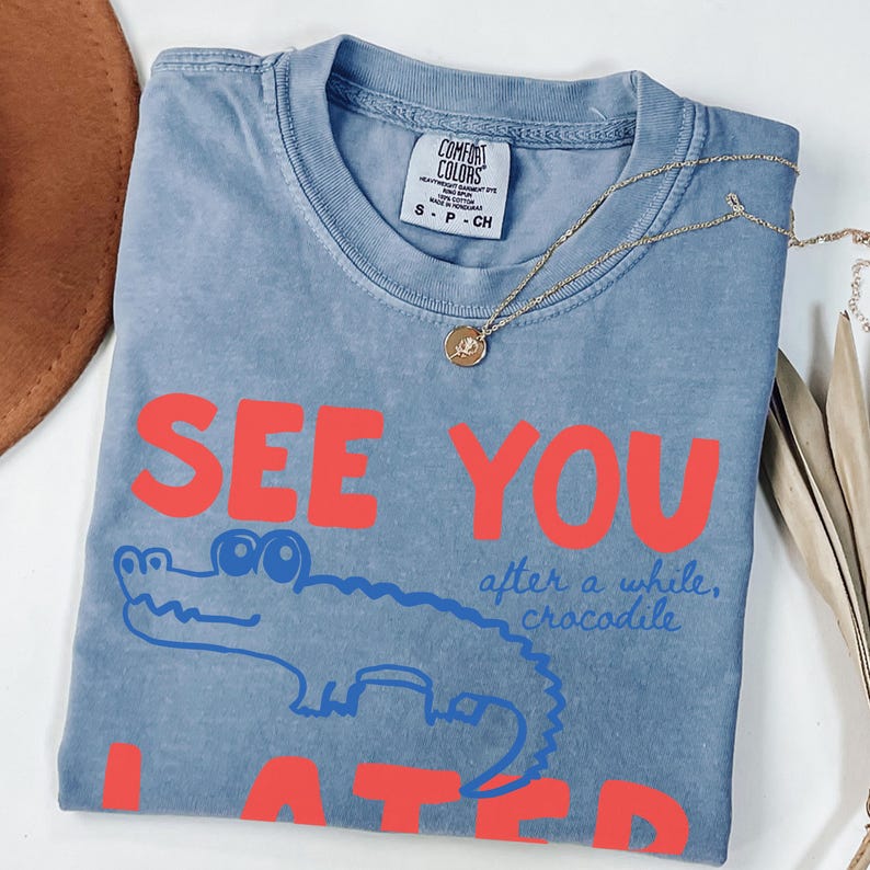 May include: A blue Comfort Colors t-shirt with the words "SEE YOU LATER" in red block letters. A blue cartoon crocodile is under the words. The phrase "after a while, crocodile" is written in blue script. A gold necklace is on the shirt.