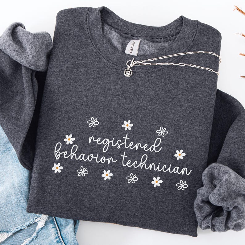 K&ouml;nnte beinhalten: Dunkelgrauer Pullover mit dem Text "registered behavior technician" in wei&szlig;er Schrift und einem G&auml;nsebl&uuml;mchenmuster um den Text herum.