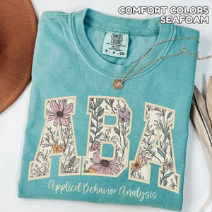 May include: Seafoam green t-shirt with the text "ABA" in a floral design. The words "Applied Behavior Analysis" are printed below the letters. A gold necklace with a pendant is draped across the shirt.