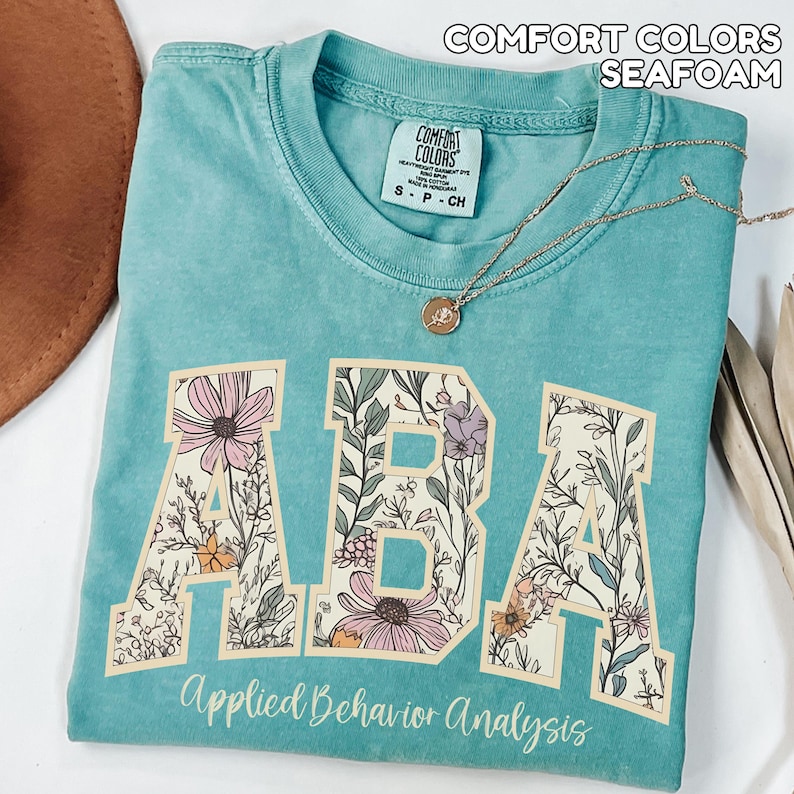 May include: Seafoam green t-shirt with the text "ABA" in a floral design. The words "Applied Behavior Analysis" are printed below the letters. A gold necklace with a pendant is draped across the shirt.