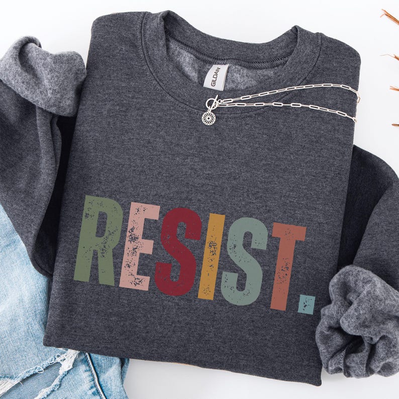 Pode incluir: Uma camisa de moletom cinza escura com a palavra "RESIST" impressa em letras coloridas e desgastadas. As letras s&atilde;o em tons de verde, rosa, vermelho, amarelo e azul.
