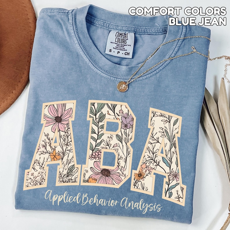 May include: A blue jean Comfort Colors t-shirt with the letters "ABA" in a floral design. The words "Applied Behavior Analysis" are printed below the letters. A gold necklace with a pendant is draped across the shirt.