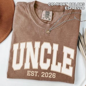 May include: Espresso-colored t-shirt with the word "UNCLE" in large, off-white block letters. Below the word is "EST. 2026". The shirt has a crew neck and a small tag. A silver necklace with a pendant is draped across the shirt.