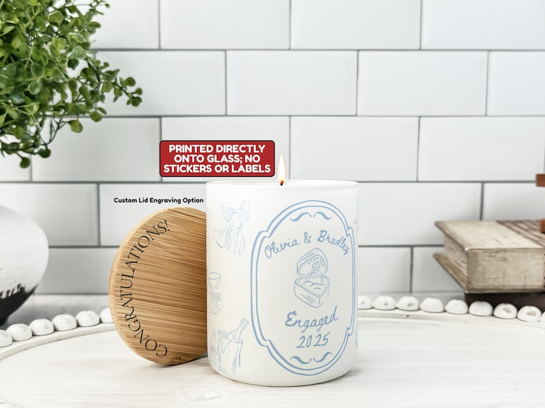 May include: A white candle with a blue design and the words "Olivia & Bradley Engaged 2025." The candle has a wooden lid with the word "CONGRATULATIONS!" printed on it. The candle is printed directly onto the glass.