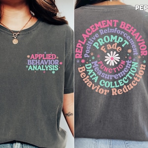 May include: A dark gray t-shirt with a graphic print on the front and back. The front print says "*APPLIED* BEHAVIOR *ANALYSIS*" in pink, blue, and green. The back print is a circle with the words "REPLACEMENT BEHAVIOR" in pink, blue, and green. The words "Positive Reinforcement", "PROMPT", "Fade", "FUNCTION", "Measurement", "DATA COLLECTION", and "Behavior Reduction" are arranged in a circle around the words "REPLACEMENT BEHAVIOR".