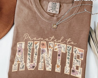 May include: An espresso brown short-sleeve t-shirt with a crew neckline. The shirt features the text "Promoted to AUNTIE" in a cream-coloured font, with the word "AUNTIE" filled with a floral pattern of pink, orange, and green flowers.