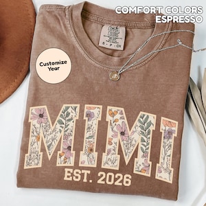 May include: Espresso-colored t-shirt with the word "MIMI" in large, floral-filled letters. The text "EST. 2026" is below. The shirt has a crew neck and a small tag. The words "COMFORT COLORS" and "ESPRESSO" are at the top.