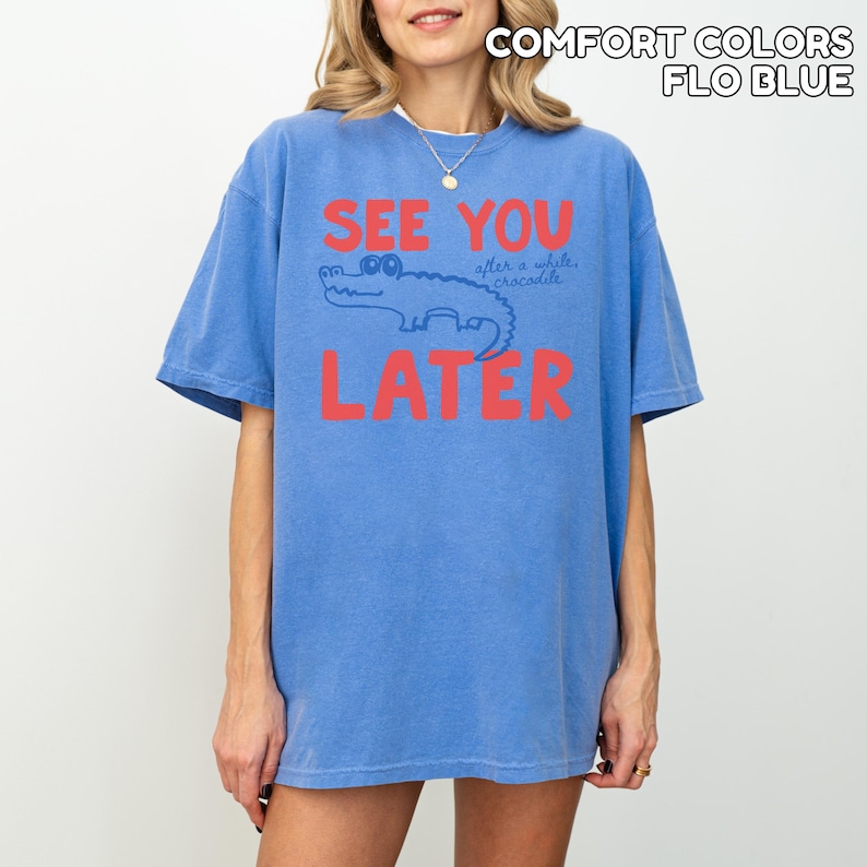 May include: A blue t-shirt with the words "SEE YOU LATER" in red block letters. A cartoon alligator is between the words. The text "after a while crocodile" is above the alligator. The shirt is labeled "COMFORT COLORS FLO BLUE".
