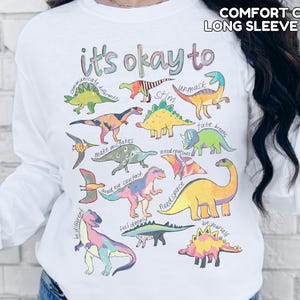 May include: White long-sleeve shirt with a colorful dinosaur graphic and the text "it's okay to" with various phrases. The shirt also has the text "COMFORT COLORS LONG SLEEVE WHITE" at the top.