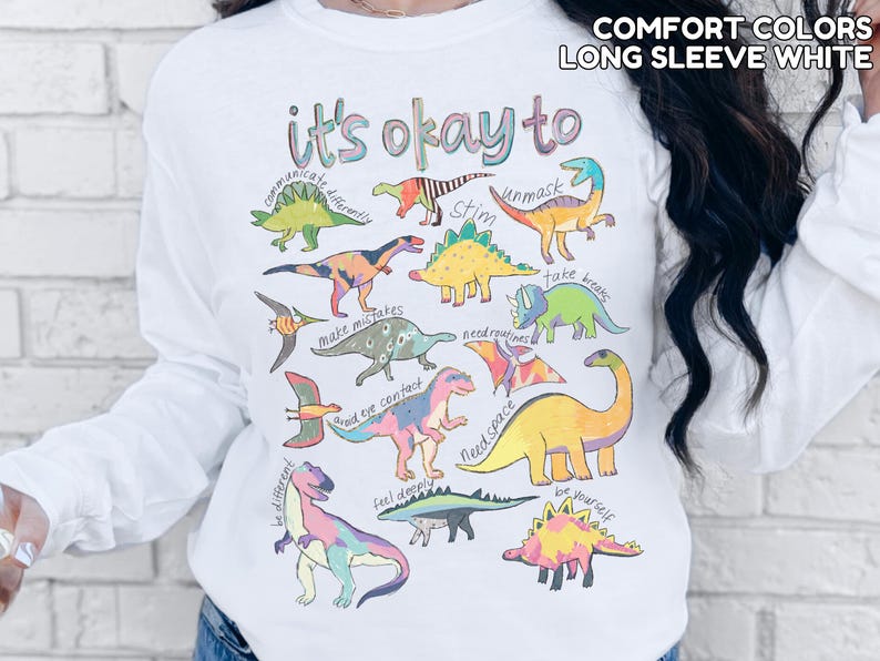 May include: White long-sleeve shirt with a colorful dinosaur graphic and the text "it's okay to" with various phrases. The shirt also has the text "COMFORT COLORS LONG SLEEVE WHITE" at the top.