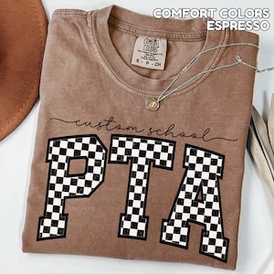 May include: Espresso-colored t-shirt with the text "custom school PTA" in black script and bold, checkered letters. The shirt features a crew neck and a small tag. A silver necklace with a pendant is draped across the shirt.