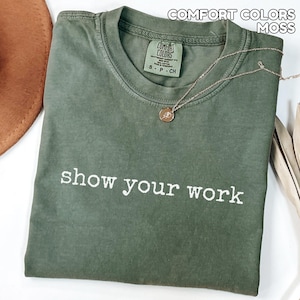 K&ouml;nnte beinhalten: Ein moosgr&uuml;nes Comfort Colors T-Shirt mit dem wei&szlig;en Schriftzug "show your work". Eine goldfarbene Halskette mit Anh&auml;nger liegt &uuml;ber dem Shirt. Das Shirt ist sauber auf einer wei&szlig;en Oberfl&auml;che gefaltet.
