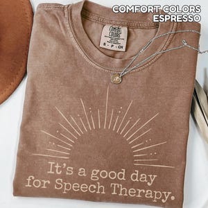 May include: Espresso-colored t-shirt with the text "It's a good day for Speech Therapy." The shirt features a sunburst design and a Comfort Colors label. A silver necklace with a pendant is draped across the shirt.