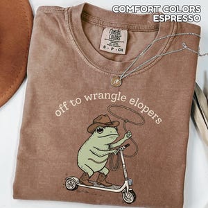 May include: Espresso-colored t-shirt with the text "off to wrangle elopers." The shirt features a cartoon frog wearing a cowboy hat and boots, riding a scooter and holding a lasso. A silver necklace is draped on the shirt.