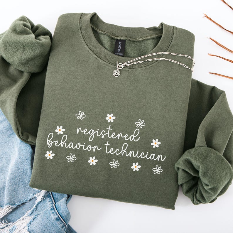 K&ouml;nnte beinhalten: Ein gr&uuml;ner Pullover mit dem Text "registered behavior technician" in wei&szlig;er Schrift und kleinen wei&szlig;en G&auml;nsebl&uuml;mchen um den Text herum.
