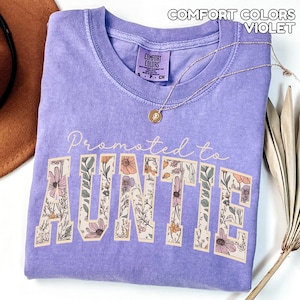 Puede incluir: Una camiseta violeta Comfort Colors con el texto "Promoted to Auntie" en un dise&ntilde;o floral. Un collar dorado con un colgante descansa sobre la camiseta. La camiseta est&aacute; doblada y un sombrero marr&oacute;n es visible en el fondo.