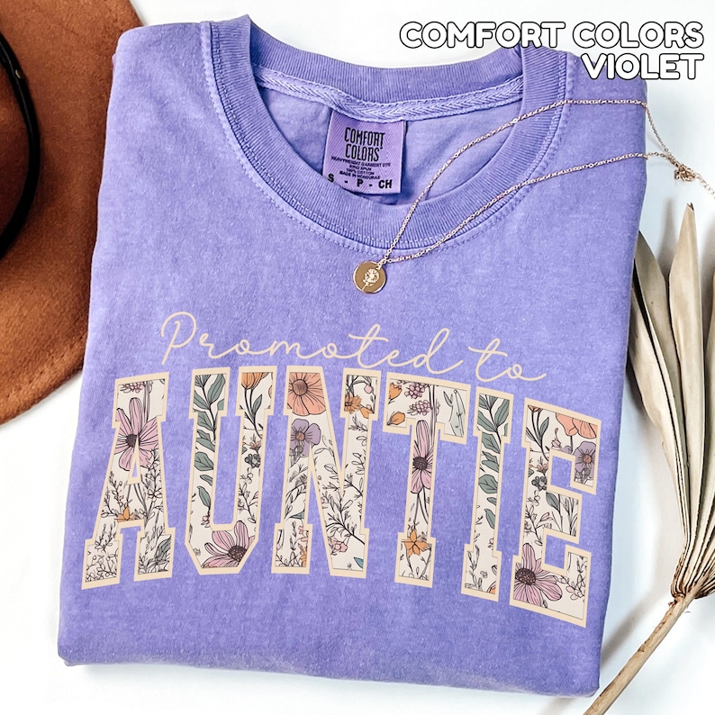 Puede incluir: Una camiseta violeta Comfort Colors con el texto "Promoted to Auntie" en un dise&ntilde;o floral. Un collar dorado con un colgante descansa sobre la camiseta. La camiseta est&aacute; doblada y un sombrero marr&oacute;n es visible en el fondo.