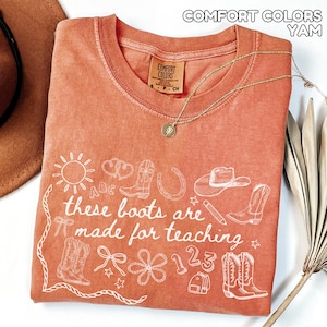 May include: A coral-colored t-shirt with a white graphic design featuring cowboy boots, a horseshoe, a hat, and the text "These boots are made for teaching." A gold necklace with a pendant is draped across the shirt.