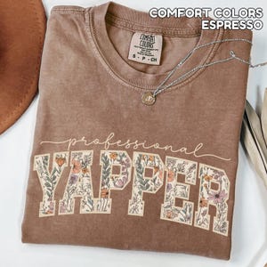 May include: Espresso-colored t-shirt with the words "professional YAPPER" in a floral design. The shirt has a crew neck and a small tag. A silver necklace with a pendant is draped across the shirt.