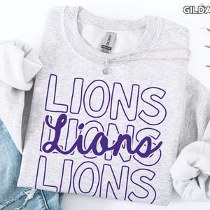 May include: A light gray sweatshirt with the word "LIONS" in a purple outlined font. The word is repeated three times, with the middle word in a cursive style. The brand name "GILDAN ASH" is visible in the top right corner.
