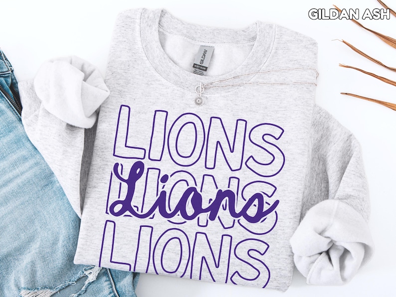 May include: A light gray sweatshirt with the word "LIONS" in a purple outlined font. The word is repeated three times, with the middle word in a cursive style. The brand name "GILDAN ASH" is visible in the top right corner.