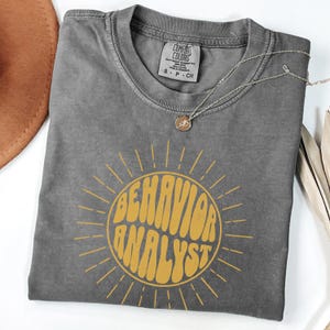 May include: A gray t-shirt with a yellow sunburst design and the text "BEHAVIOR ANALYST" in a retro font.