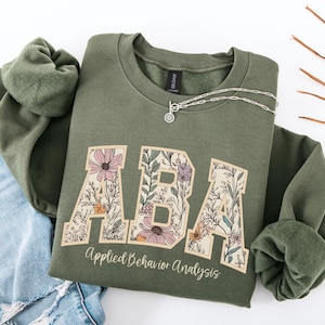 May include: An olive green sweatshirt featuring the letters "ABA" in a floral design. The words "Applied Behavior Analysis" are printed below. A silver necklace with a pendant is draped on the sweatshirt.