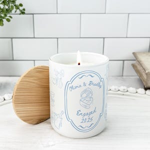 May include: A white candle with a wooden lid, lit with a flame. The candle has a blue design with the words "Olivia & Bradley" and "Engaged 2025". The candle is on a white tray.