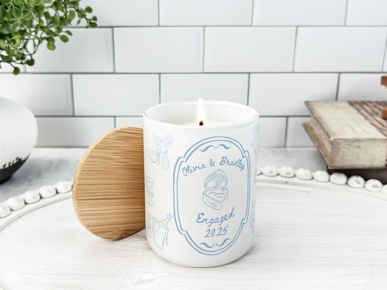 May include: A white candle with a wooden lid, lit with a flame. The candle has a blue design with the words "Olivia & Bradley" and "Engaged 2025". The candle is on a white tray.