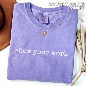 K&ouml;nnte beinhalten: Ein violettes Comfort Colors T-Shirt mit dem wei&szlig;en Schriftzug "show your work". Eine goldene Halskette mit Anh&auml;nger liegt auf dem gefalteten Shirt. Ein brauner Hut ist teilweise sichtbar.
