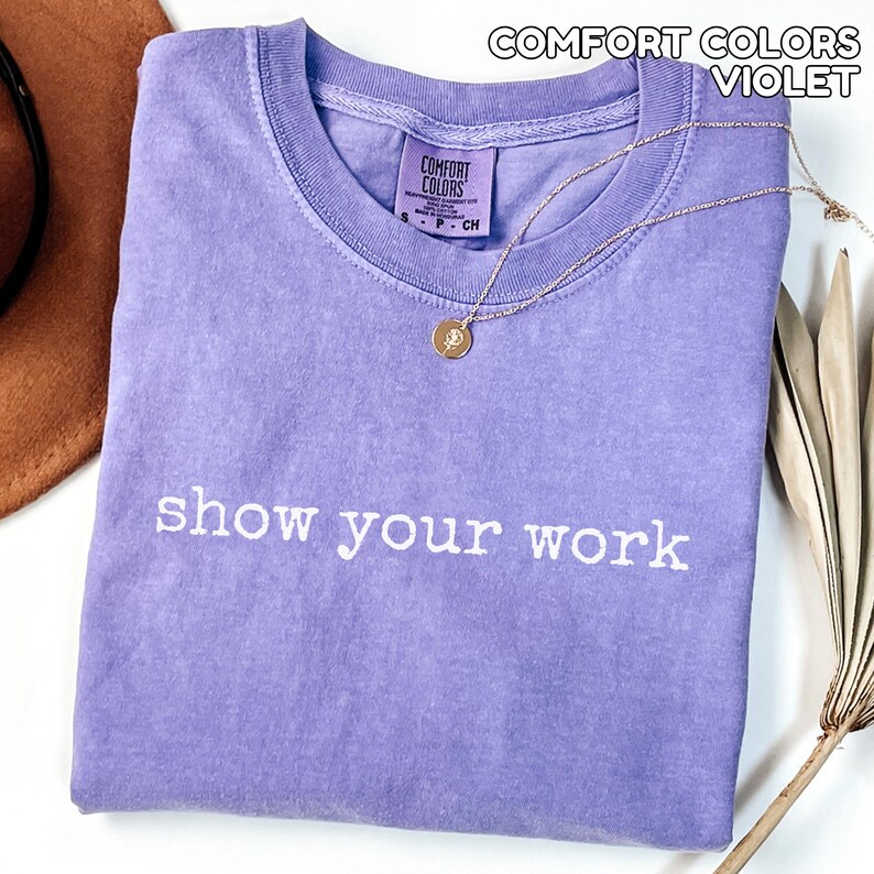 K&ouml;nnte beinhalten: Ein violettes Comfort Colors T-Shirt mit dem wei&szlig;en Schriftzug "show your work". Eine goldene Halskette mit Anh&auml;nger liegt auf dem gefalteten Shirt. Ein brauner Hut ist teilweise sichtbar.