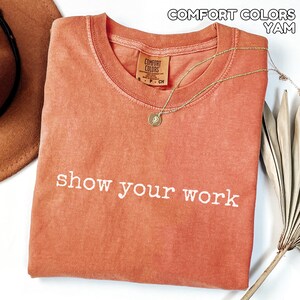 K&ouml;nnte beinhalten: Ein korallenfarbenes T-Shirt mit dem wei&szlig;en Schriftzug "show your work". Das Shirt hat einen Rundhalsausschnitt und ein kleines Comfort Colors-Etikett. Eine goldene Halskette mit Anh&auml;nger liegt auf dem Shirt.