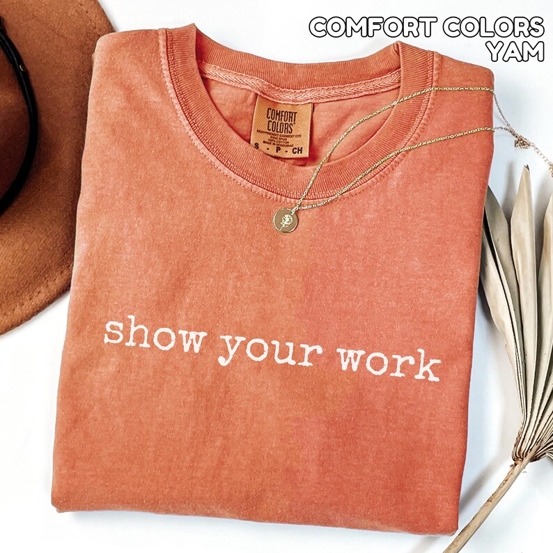 K&ouml;nnte beinhalten: Ein korallenfarbenes T-Shirt mit dem wei&szlig;en Schriftzug "show your work". Das Shirt hat einen Rundhalsausschnitt und ein kleines Comfort Colors-Etikett. Eine goldene Halskette mit Anh&auml;nger liegt auf dem Shirt.