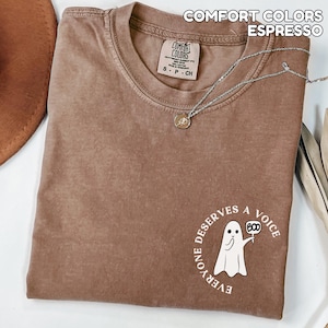 May include: Espresso-colored Comfort Colors t-shirt with a round neck and short sleeves. The shirt features a graphic of a ghost holding a sign that says "BOO" and the text "EVERYONE DESERVES A VOICE". A silver necklace with a pendant is draped across the shirt.