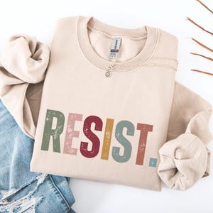 Pode incluir: Uma camisa de moletom bege com a palavra "RESIST" impressa em letras coloridas e desgastadas. As letras s&atilde;o em tons de verde, rosa, vermelho, amarelo e azul.