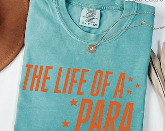 May include: Seafoam green t-shirt with the text "THE LIFE OF A PARA" in orange, with orange star accents. The shirt has a crew neck and short sleeves. A gold necklace with a pendant is draped across the shirt.