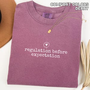 May include: A berry-coloured Comfort Colors t-shirt with the phrase "regulation before expectation" printed in white. A gold necklace with a pendant rests on the shirt. The shirt is folded and displayed with a hat and dried plant.