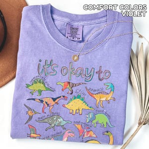 May include: A violet Comfort Colors t-shirt with the phrase "it's okay to" and colorful dinosaur illustrations. The shirt also features the words "communicate differently", "stim", "unmask", "make mistakes", "avoid eye contact", "need routines", and "take breaks". A gold necklace is draped across the shirt.