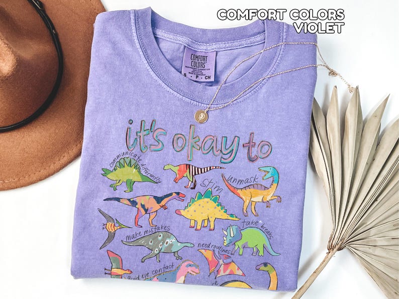 May include: A violet Comfort Colors t-shirt with the phrase "it's okay to" and colorful dinosaur illustrations. The shirt also features the words "communicate differently", "stim", "unmask", "make mistakes", "avoid eye contact", "need routines", and "take breaks". A gold necklace is draped across the shirt.
