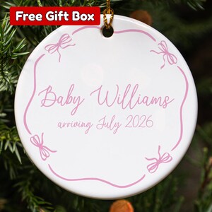 May include: A white ceramic Christmas ornament with pink text and bow accents. The ornament reads "Baby Williams arriving July 2026". The ornament is hanging on a Christmas tree. A red box with the text "Free Gift Box" is in the top left corner.