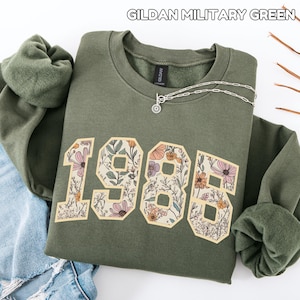 Op de afbeelding: Een olijfgroene sweatshirt met de tekst "1985" in een bloemenontwerp. De sweatshirt heeft een ronde hals en lange mouwen. Een zilveren ketting met een hanger is over de voorkant gedrapeerd. De tekst "GILDAN MILITARY GREEN" staat bovenaan.