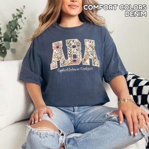 May include: A denim blue t-shirt with the letters "ABA" in a floral design. The words "Applied Behavior Analysis" are below the letters. The shirt is part of the Comfort Colors collection.