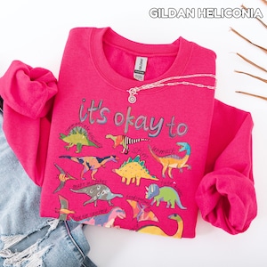 May include: A bright pink sweatshirt with the text "it's okay to" and various dinosaur illustrations. The sweatshirt is paired with a pair of blue jeans with distressed details and a silver necklace.