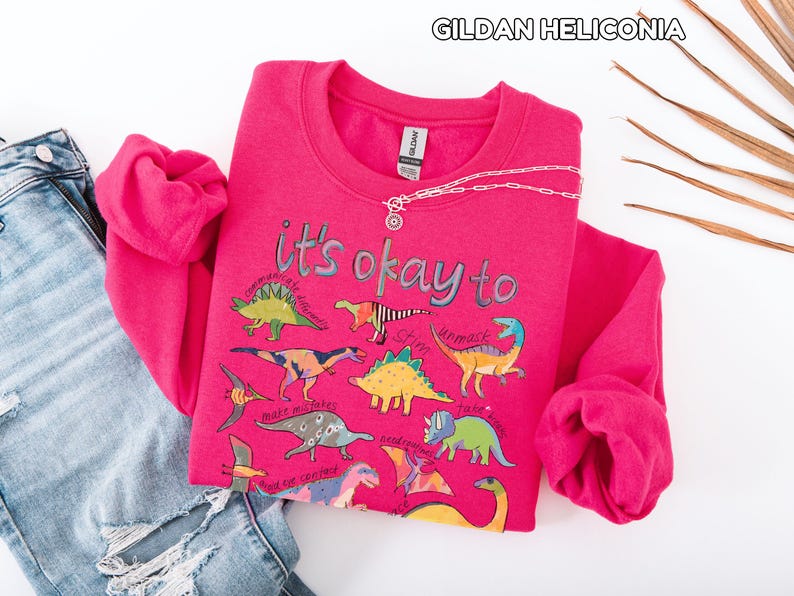 May include: A bright pink sweatshirt with the text "it's okay to" and various dinosaur illustrations. The sweatshirt is paired with a pair of blue jeans with distressed details and a silver necklace.